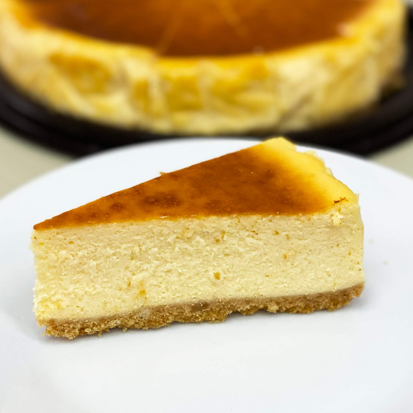 ชีสเค้กคลาสสิกหนึ่งชิ้น เสิร์ฟบนจานสีขาว พร้อมชีสเค้กทั้งก้อนอยู่ด้านหลัง